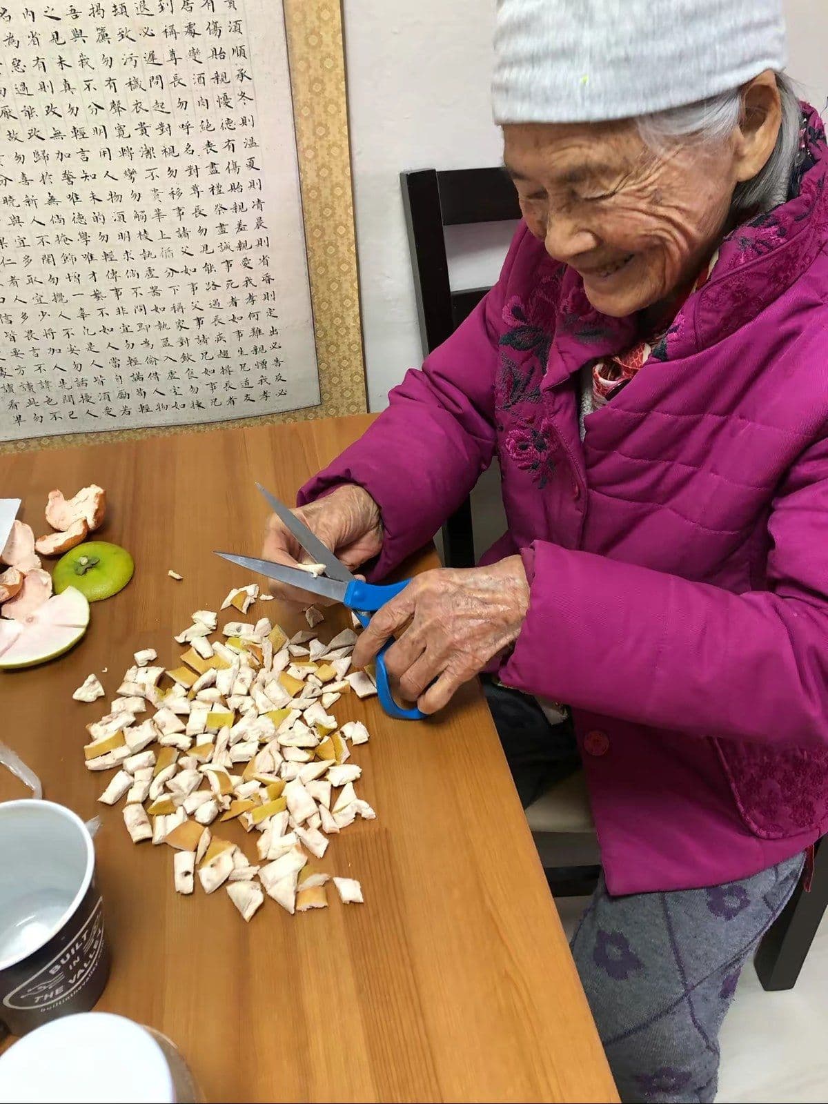 Our mother, 99, smiling as she cuts pomelo peel into small pieces to dry at the kitchen table.