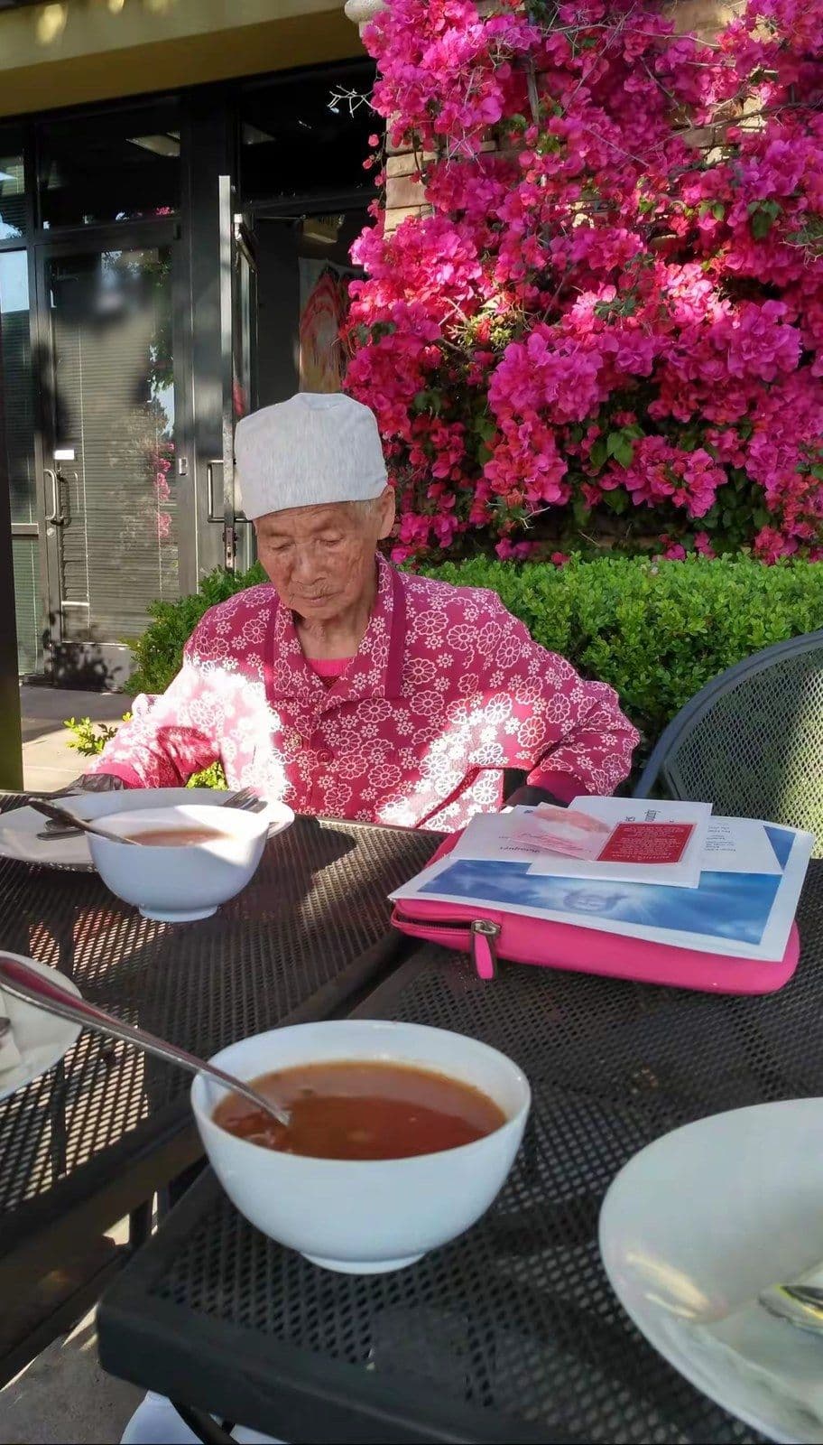 Our mother, 99, sitting down to a bowl of soup at an outdoor table beside bright pink flowers.